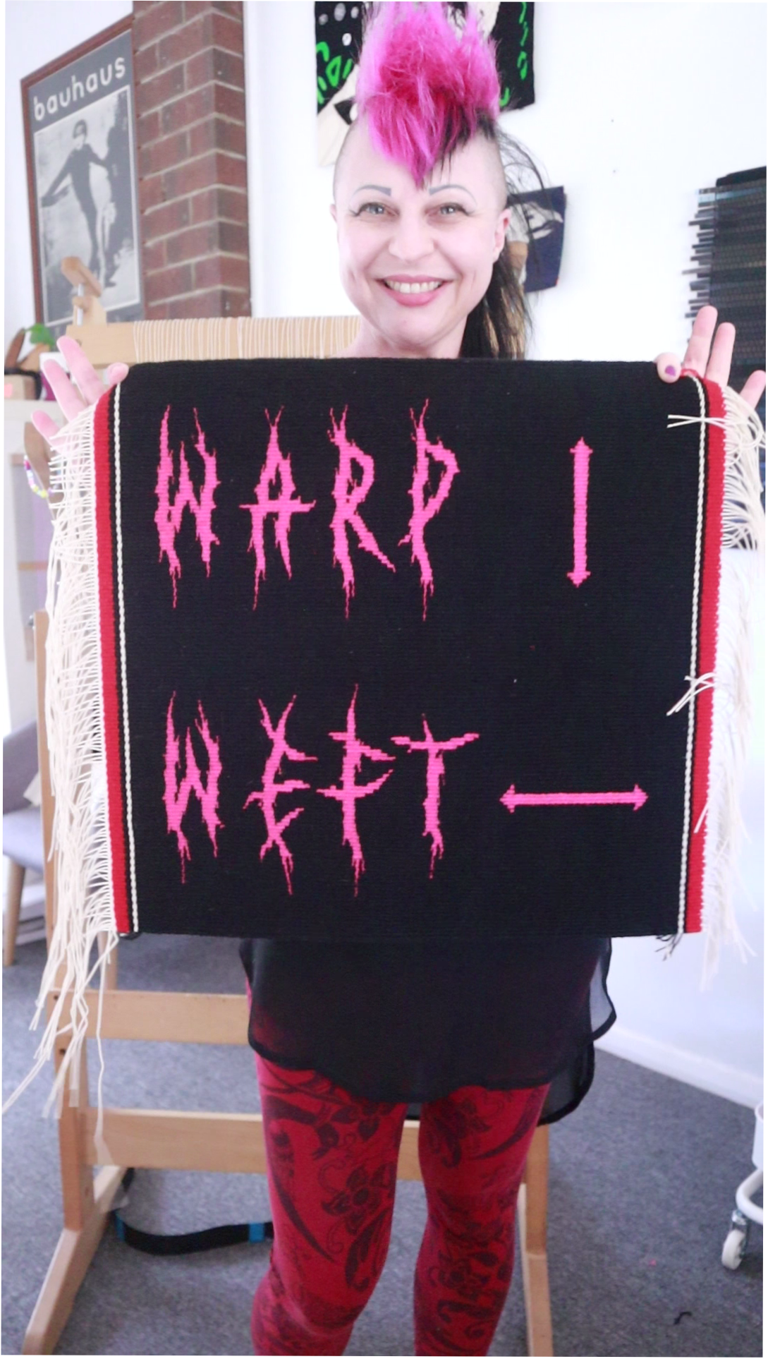 Michelle Driver with her tapestry - Artwork made in handwoven tapestry with the words warp and weft in pink deathmetal style lettering